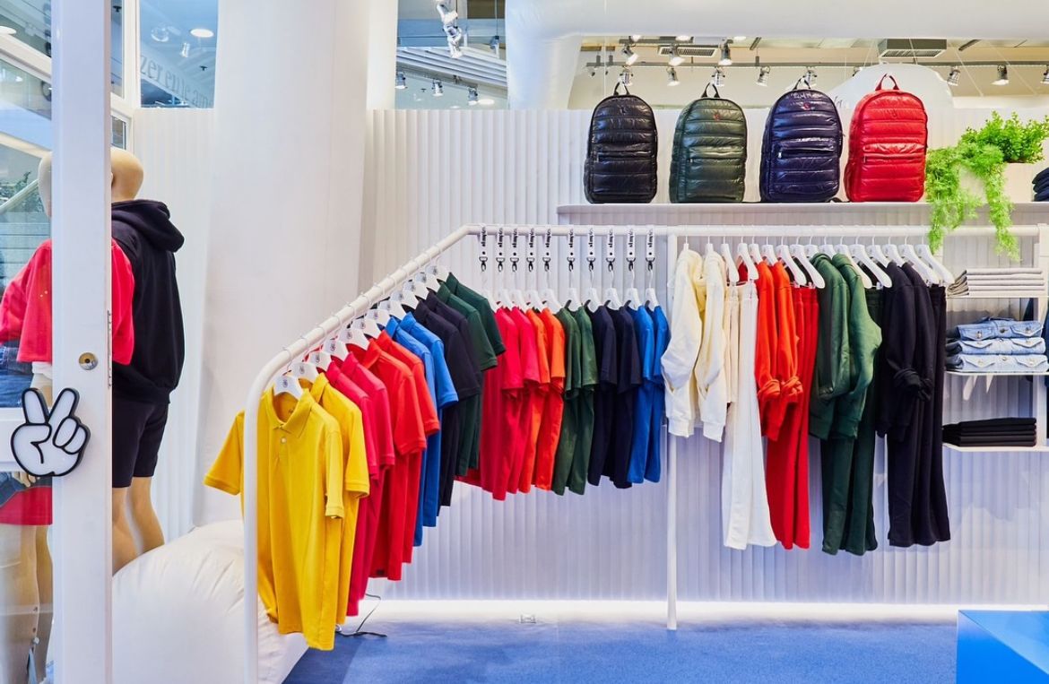 Parede modular com arara branca de polos coloridas em degradê e expositor superior de mochilas