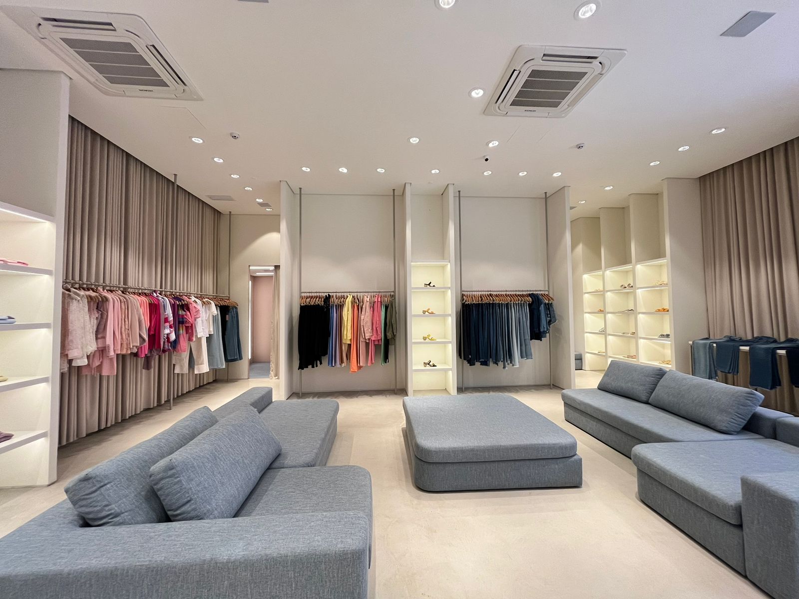 Salão principal da sede Carol com ilhas de sofás cinza, cortinas de linho e expositores modulares brancos