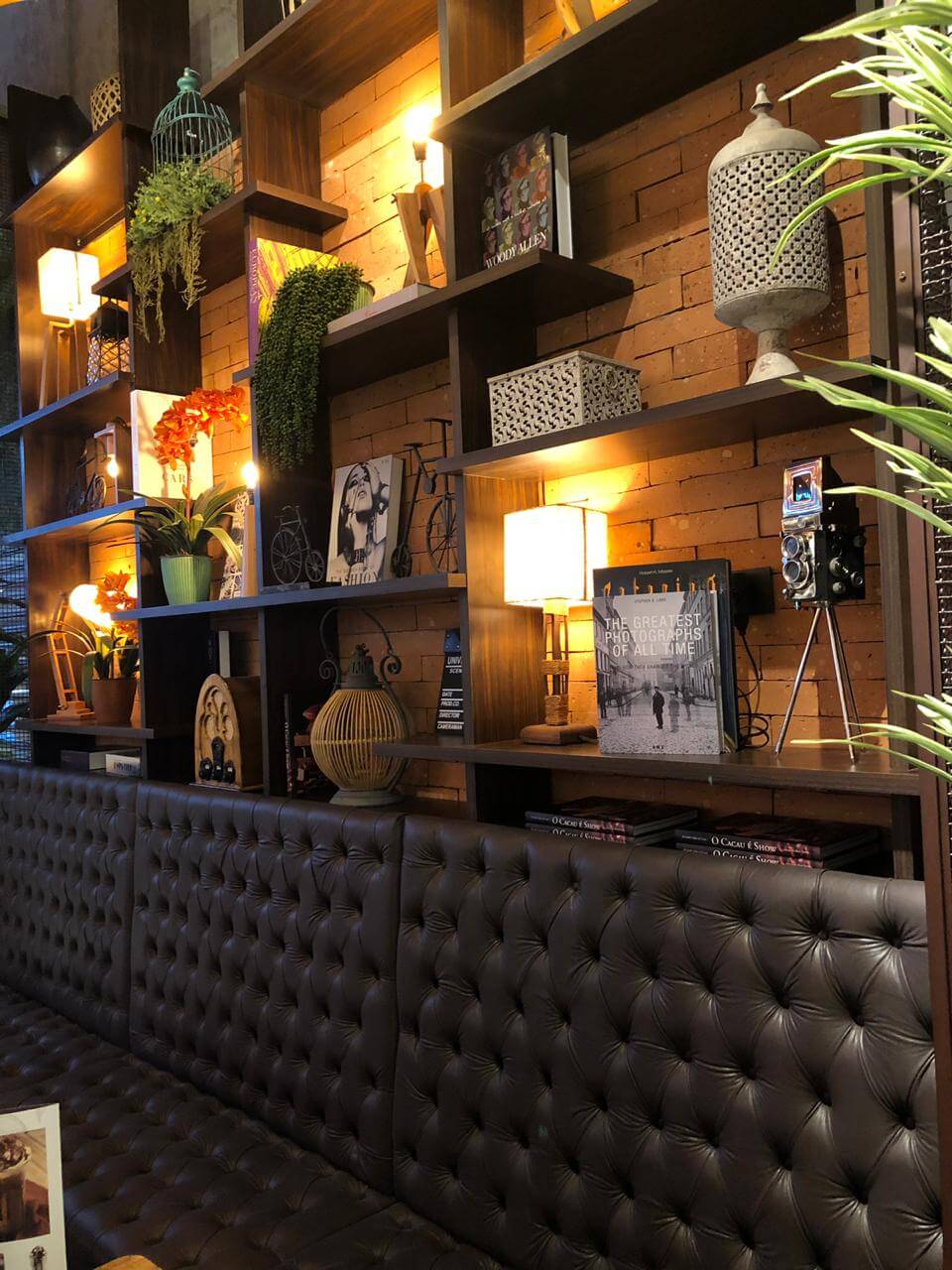 Área lounge da cafeteria com biblioteca cenográfica em tijolinho aparente, estante de madeira e sofá chesterfield de couro